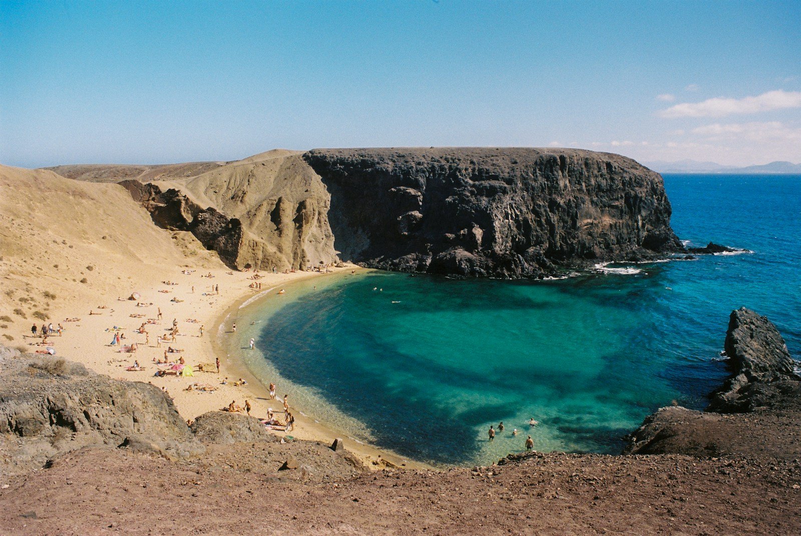 a sandy beach with clear blue water next to a cliff